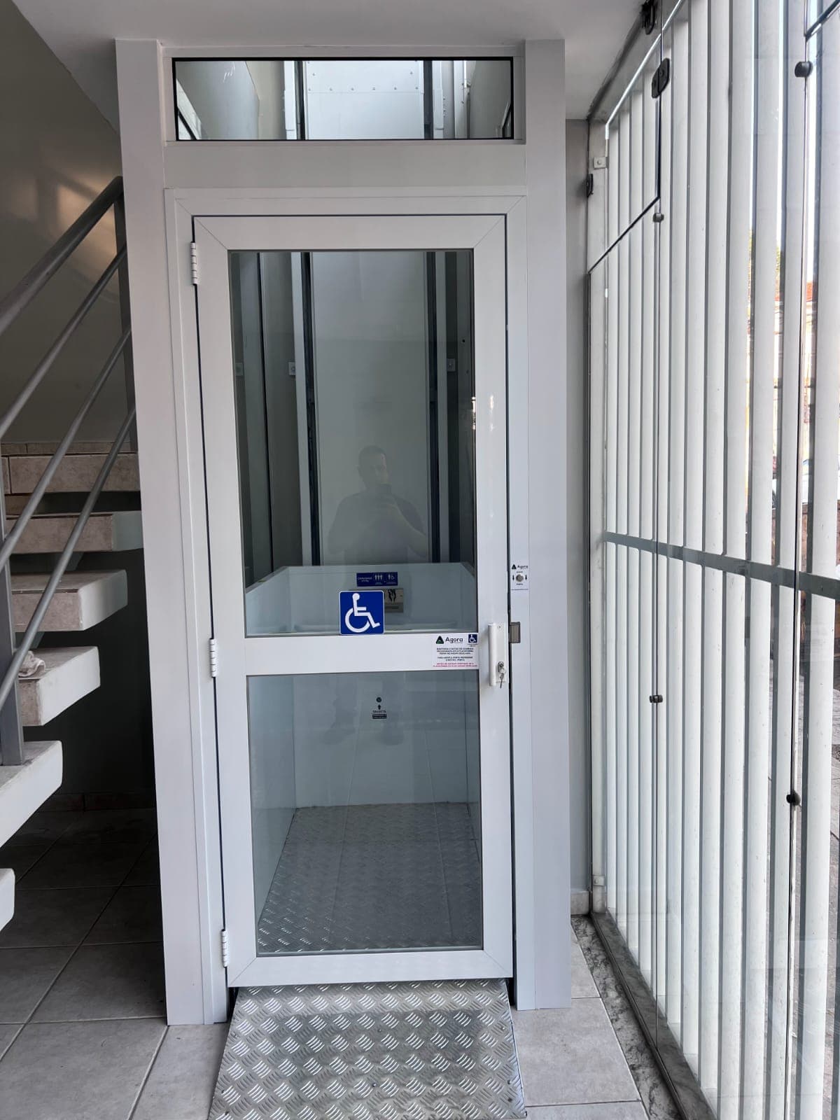 Elevador hidráulico enclausurado em estrutura branca, instalado junto à escada externa